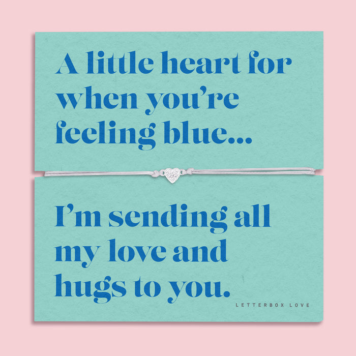 A teal-colored bracelet card titled "Feeling Blue" from Letterbox Love, featuring a small heart charm on a grey string bracelet. The card includes the message: "A little heart for when you’re feeling blue... I’m sending all my love and hugs to you." The setting is a soft pink background with small blue flowers and decorative gems scattered around.