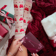 A person's legs and hands are shown wearing a pair of cream-colored crew socks with red bow and heart patterns, styled with a holiday greeting card, a burgundy knit blanket, and wrapped gifts on a white wooden floor.
