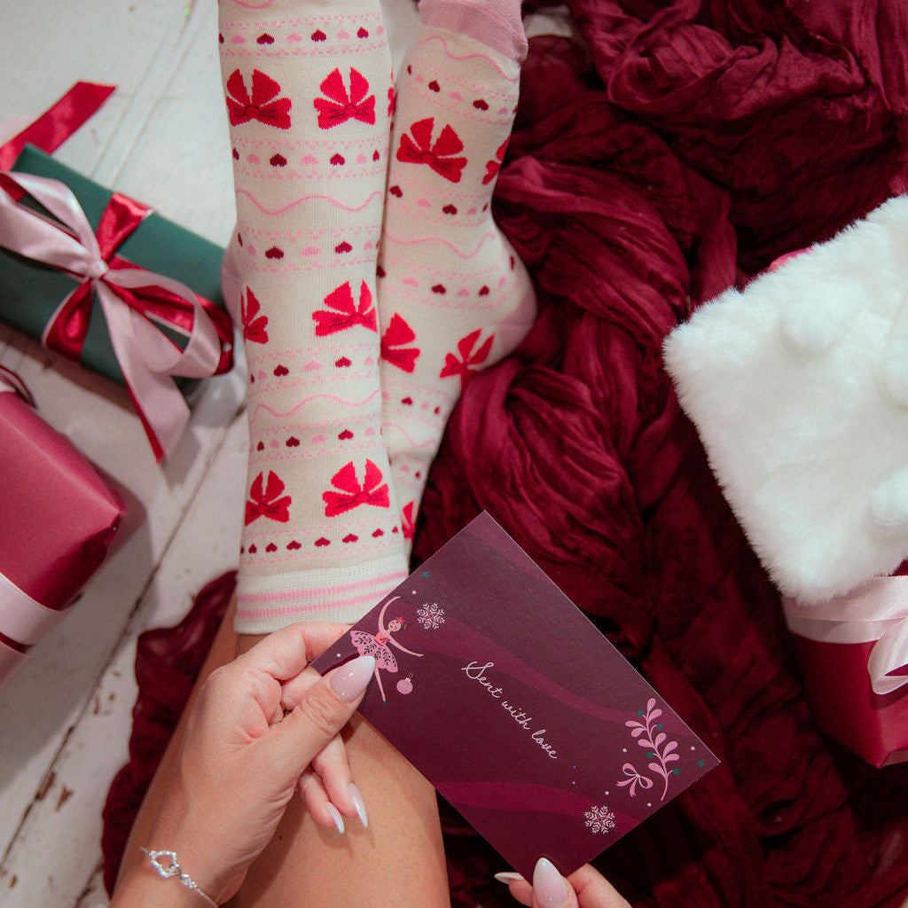 A person's legs and hands are shown wearing a pair of cream-colored crew socks with red bow and heart patterns, styled with a holiday greeting card, a burgundy knit blanket, and wrapped gifts on a white wooden floor.