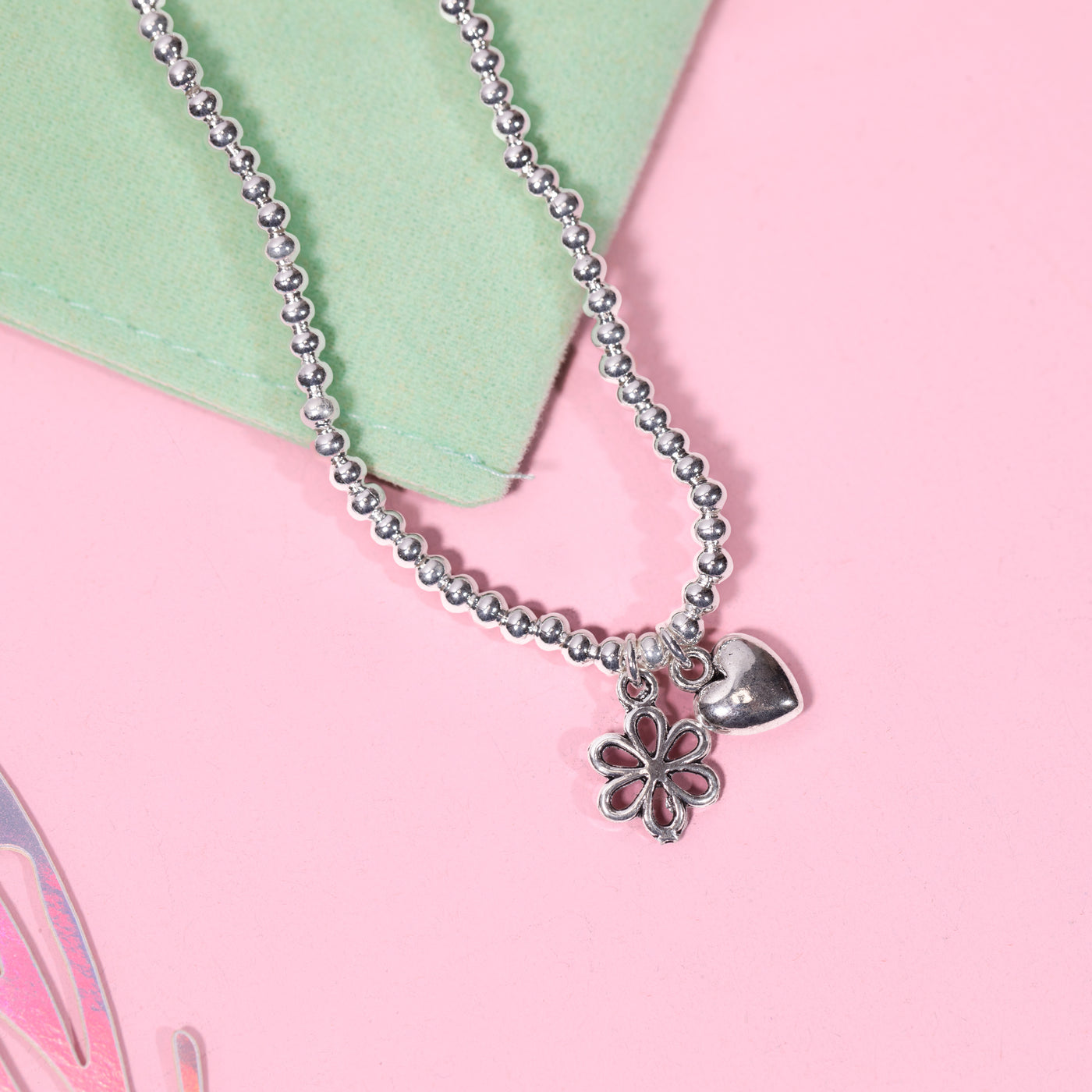A silver-plated beaded bracelet with a flower and heart charm for children, displayed on a pink background with a light green velvet gift wrap