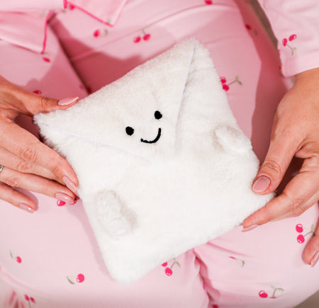 Soft white fluffy envelope pouch with a cute embroidered smiley face, held in someone’s hands while wearing pink cherry-print pyjamas. A cosy gift pouch designed to hold any Letterbox Love bracelet, including boxed bracelets.