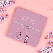 A lilac adjustable bracelet featuring two interlinked heart charms, displayed on a lilac backing card with the playful message "For Fox Sake, You’re Leaving?!" accompanied by cute fox illustrations. Surrounded by soft pink flowers. Perfect as a farewell gift.