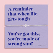 A lavender-colored bracelet card titled "Strong Stuff" from Letterbox Love, featuring a small star charm on a grey string bracelet. The card includes the message: "A reminder that when life gets tough, you’ve got this, you’re made of strong stuff." The setting is a soft pink background with small purple flowers and decorative gems scattered around.