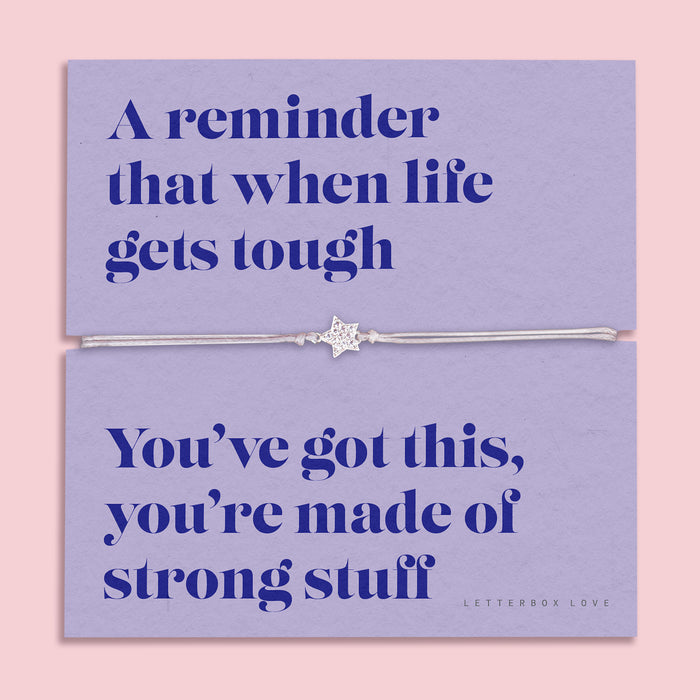 A lavender-colored bracelet card titled "Strong Stuff" from Letterbox Love, featuring a small star charm on a grey string bracelet. The card includes the message: "A reminder that when life gets tough, you’ve got this, you’re made of strong stuff." The setting is a soft pink background with small purple flowers and decorative gems scattered around.