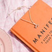 bracelet with star and moon charms on an orange book titled 'Manifest' on a pink surface. A bracelet to say well done.