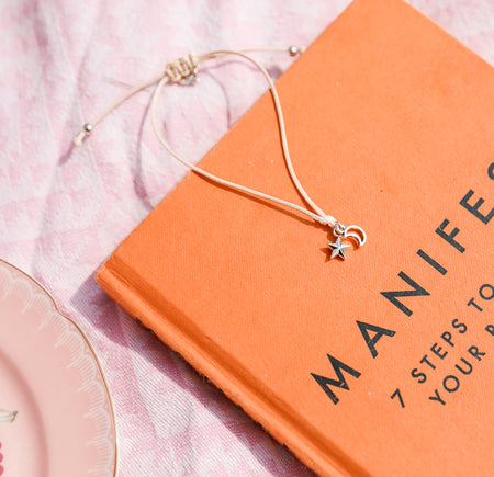 bracelet with star and moon charms on an orange book titled 'Manifest' on a pink surface. A bracelet to say well done.