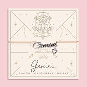 Adjustable peach cord bracelet with a silver 'Gemini' charm and heart pendant, displayed on a zodiac-themed card with a twin illustration and the traits "Playful, Spontaneous, Curious".