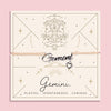 Adjustable peach cord bracelet with a silver 'Gemini' charm and heart pendant, displayed on a zodiac-themed card with a twin illustration and the traits 