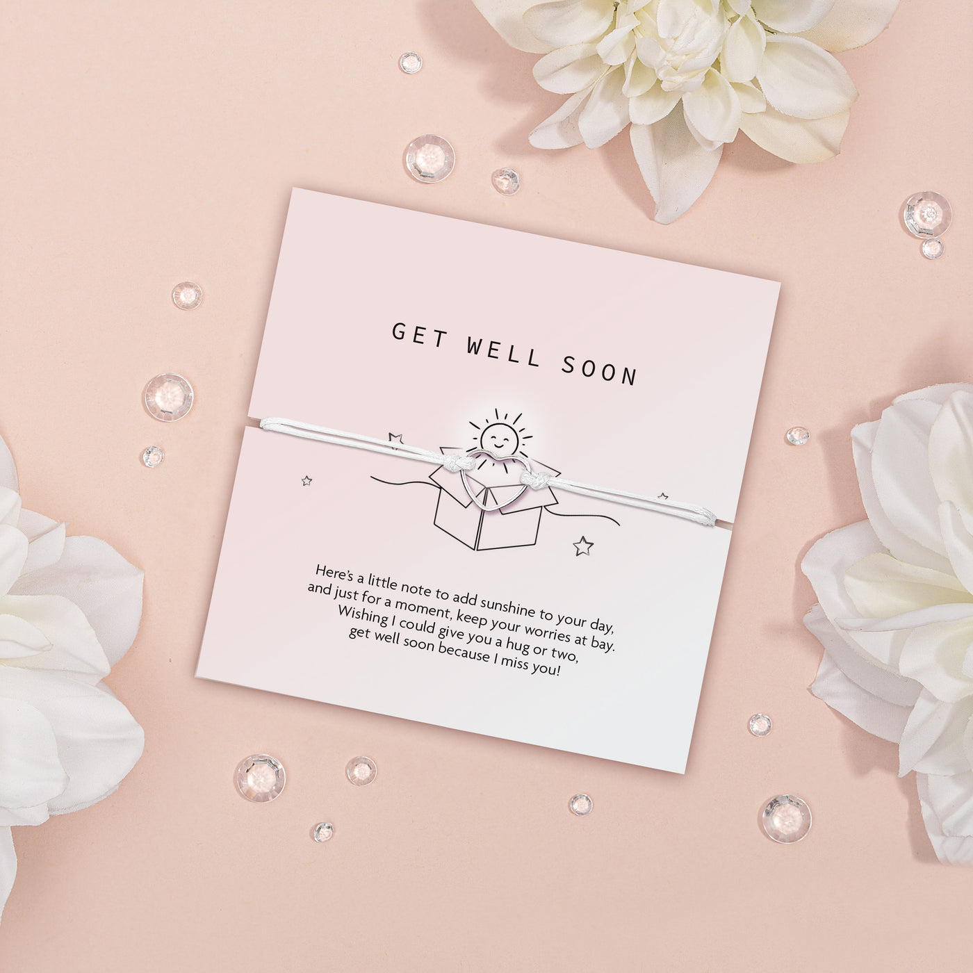 'Get Well Soon' card on a soft pink background, tied with a delicate white ribbon. The card features a cheerful sun illustration and a comforting message. Beside the card are vibrant yellow flowers with white petals and scattered clear gemstones, evoking a sense of warmth and well-wishes.