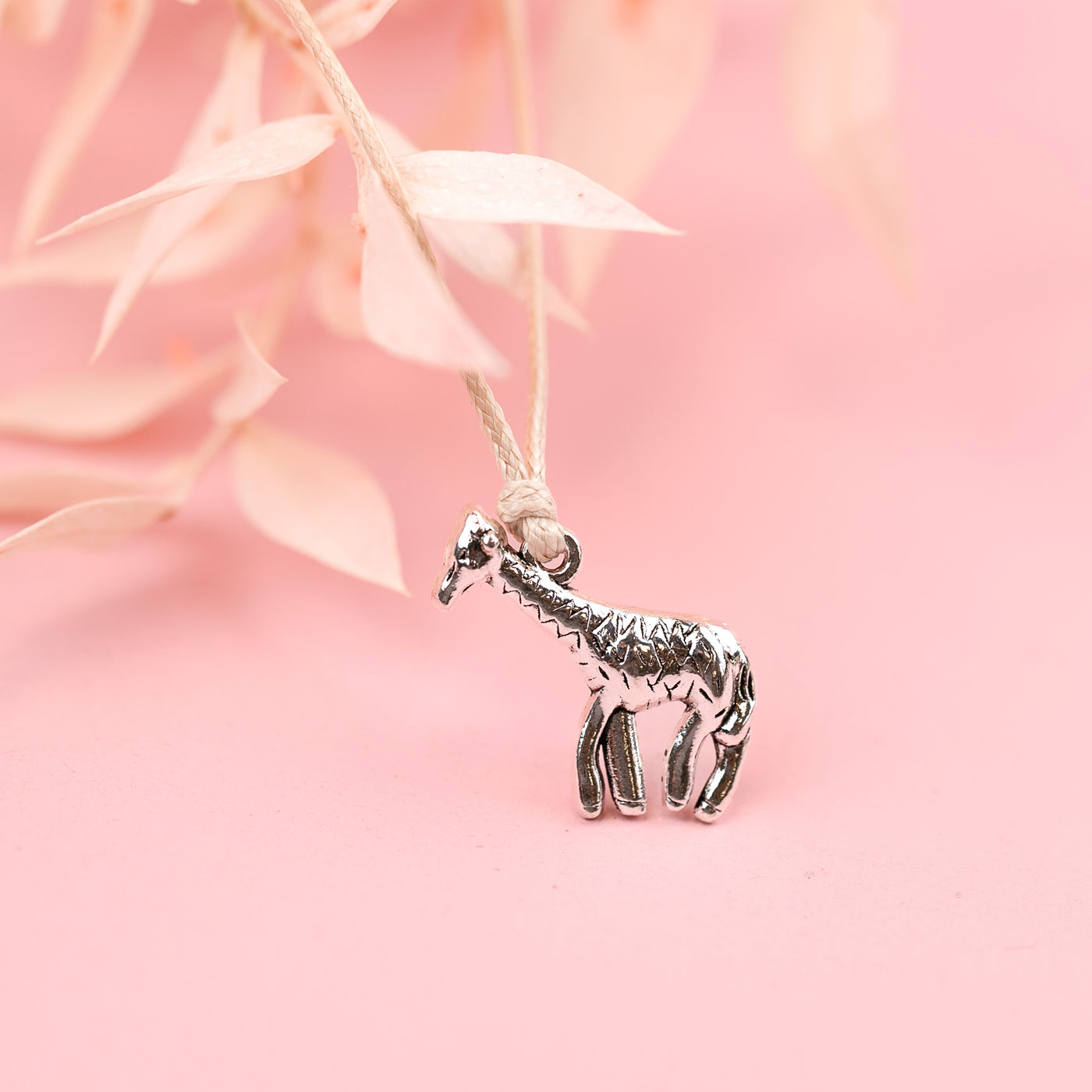 A cute silver-plated giraffe charm on a beige string friendship bracelet, with a pale pink background.