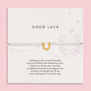 Charming 'GOOD LUCK' card on a soft pink background, adorned with encouraging words and a horseshoe illustration, accompanied by a slender white bracelet with a golden horseshoe charm.