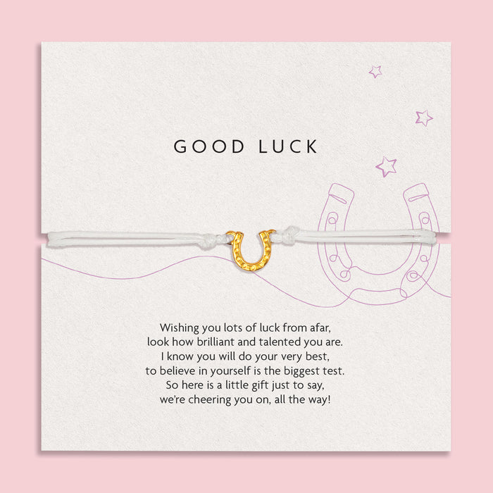 Charming 'GOOD LUCK' card on a soft pink background, adorned with encouraging words and a horseshoe illustration, accompanied by a slender white bracelet with a golden horseshoe charm.