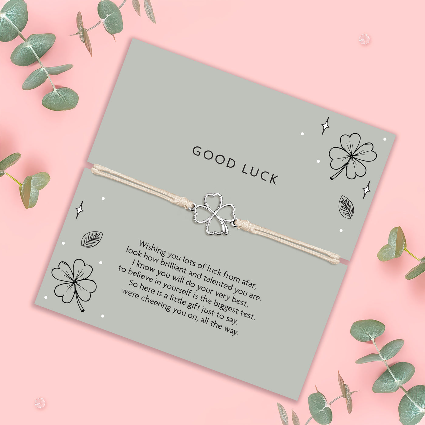 Silver four-leaf clover bracelet on a 'Good Luck' card with decorative elements on a pink background.