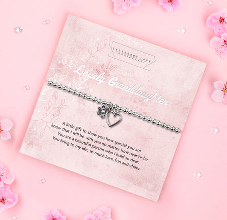 A silver beaded bracelet with a heart and flower charm displayed on a card reading 