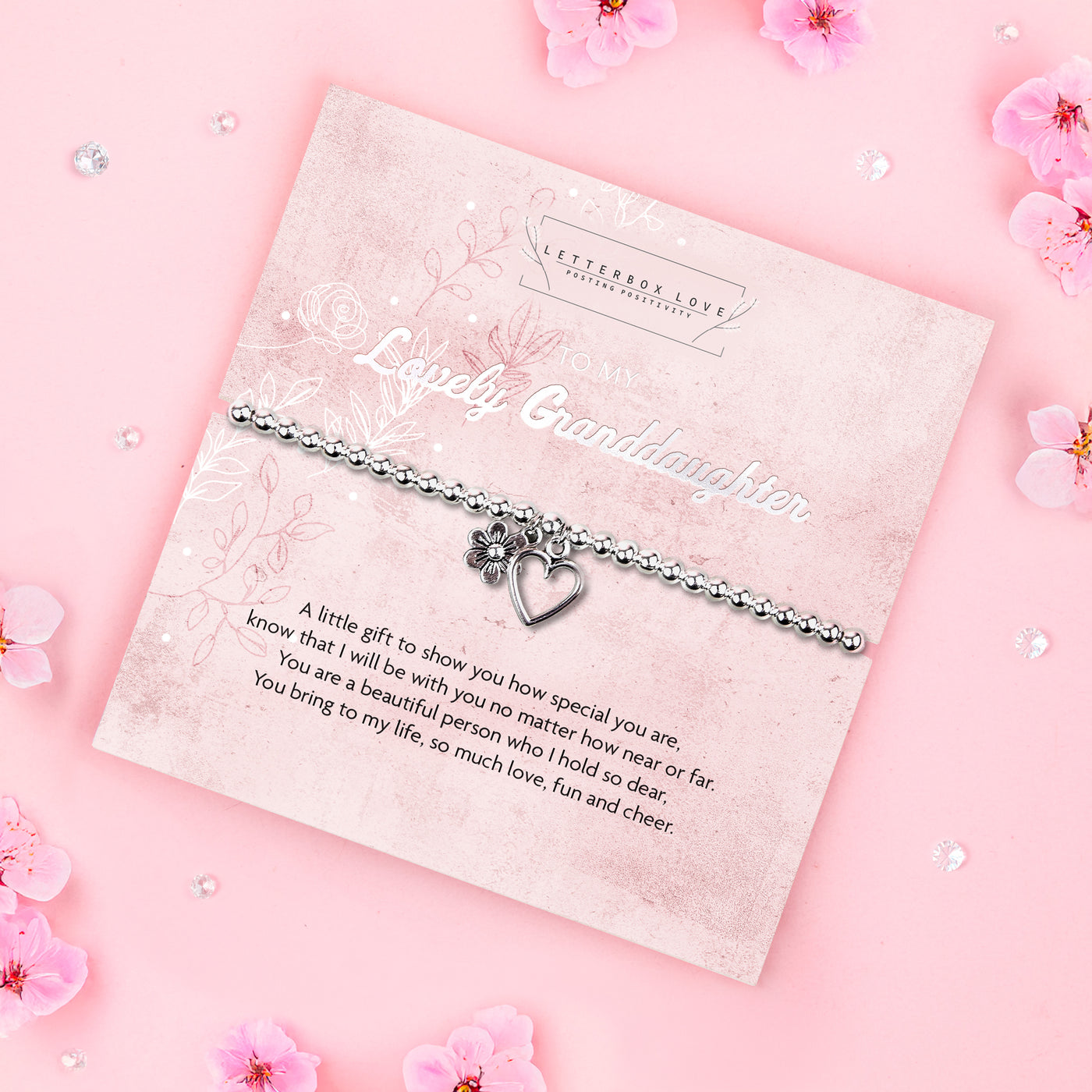 A silver beaded bracelet with a heart and flower charm displayed on a card reading 