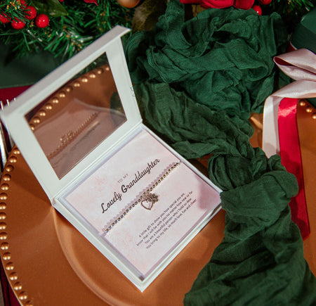 An open white jewelry gift box containing a delicate chain bracelet with a small heart & flower pendant, displayed on a pink card. The box sits on a copper-toned plate surrounded by dark green fabric, red ribbon, and holiday greenery with red berries.