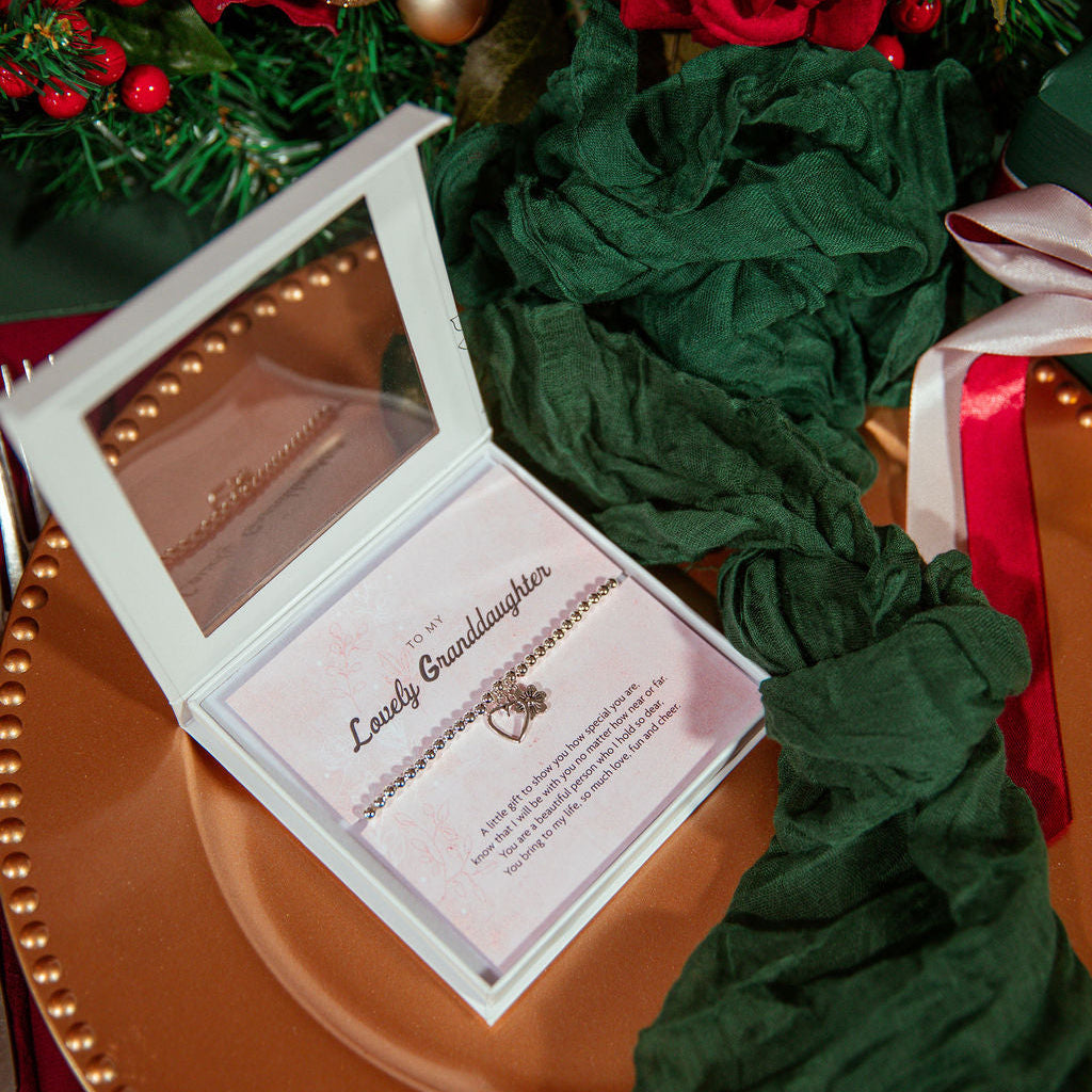 An open white jewelry gift box containing a delicate chain bracelet with a small heart & flower pendant, displayed on a pink card. The box sits on a copper-toned plate surrounded by dark green fabric, red ribbon, and holiday greenery with red berries.