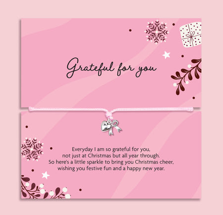 A pink Christmas-themed gift with a top card reading 'Grateful for you' and a bottom card with a message of gratitude, with an adjustable pink cord bracelet and a small silver heart and bow charm. The card features decorative snowflakes, stars, leaves, and a gift box illustration on a soft pink background.