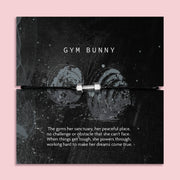A black cord bracelet with a silver plated charm shaped like a dumbbell, presented on a black backing card with the text 'GYM BUNNY' and a motivational quote, set against a solid pink background.