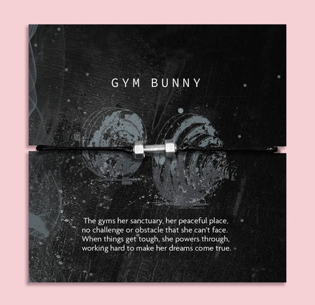 A black cord bracelet with a silver plated charm shaped like a dumbbell, presented on a black backing card with the text 'GYM BUNNY' and a motivational quote, set against a solid pink background.