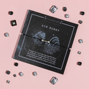 Flat lay of a 'GYM BUNNY' card from Letterbox Love on a pink background, featuring a descriptive passage about a dedicated gym-goer. The card is adorned with a thin bracelet with a square charm and is surrounded by scattered crystal gemstones of various shapes and sizes.