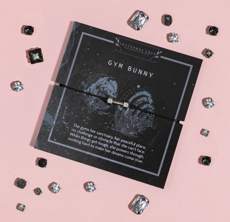 Flat lay of a 'GYM BUNNY' card from Letterbox Love on a pink background, featuring a descriptive passage about a dedicated gym-goer. The card is adorned with a thin bracelet with a square charm and is surrounded by scattered crystal gemstones of various shapes and sizes.