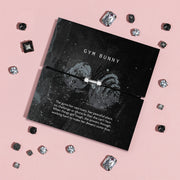 A black cord bracelet with a silver-plated dumbbell charm, displayed on a black branded card that reads 'GYM BUNNY' with inspirational text, arranged on a pink background with scattered rhinestones.