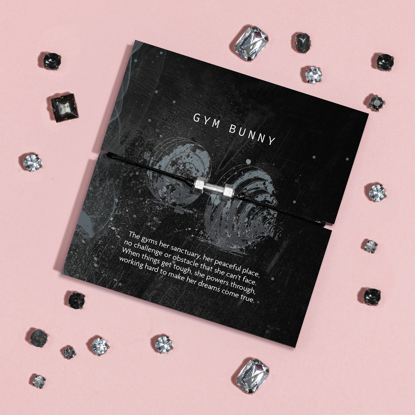 A black cord bracelet with a silver-plated dumbbell charm, displayed on a black branded card that reads 'GYM BUNNY' with inspirational text, arranged on a pink background with scattered rhinestones.