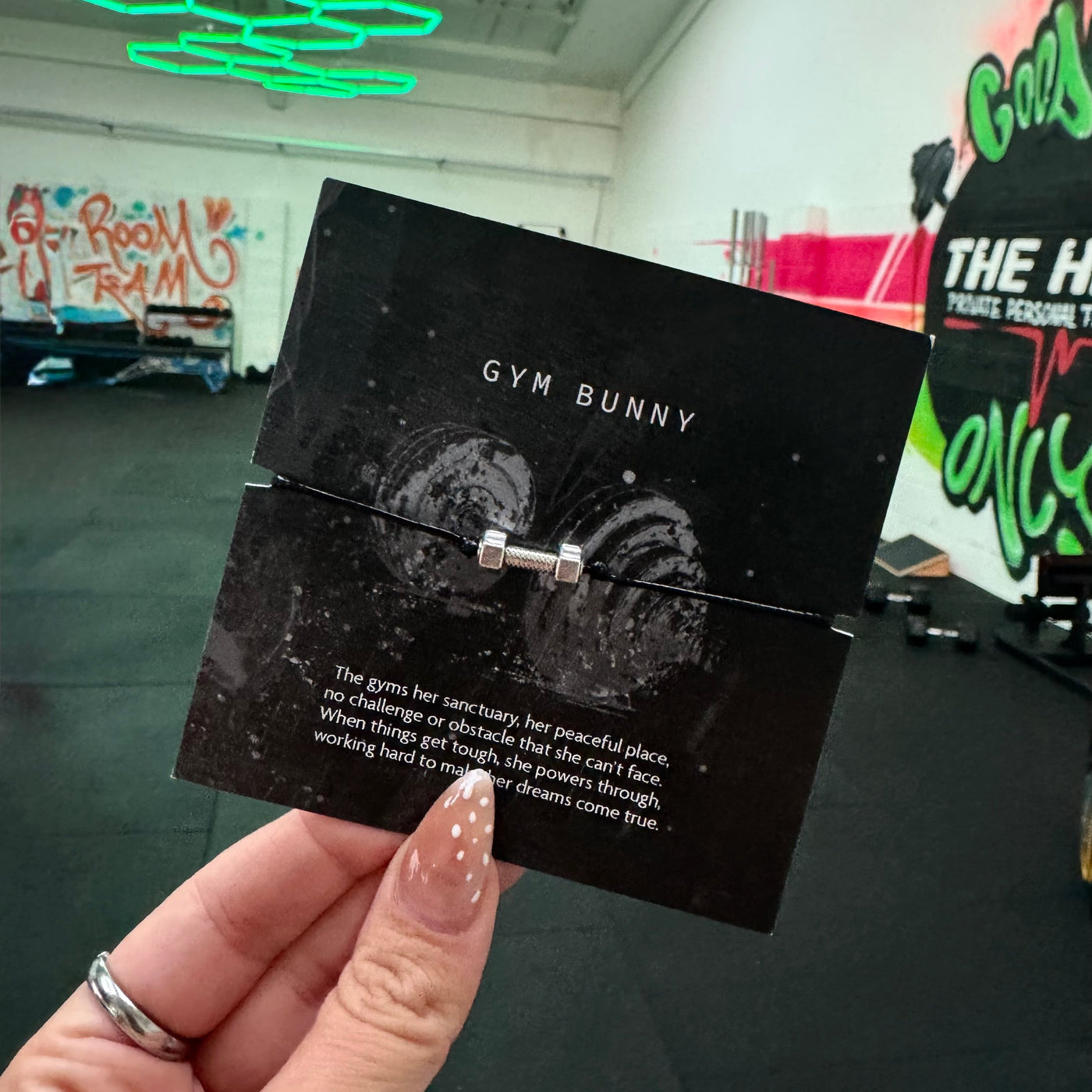 A hand holding a black card display with a small silver ring featuring a barbell-like center, printed with the words 'GYM BUNNY' and a quote. The background shows a gym interior with colorful wall art and bright ceiling lights.
