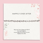 A delicate silver chain bracelet with clear rhinestones displayed on a white card with pink floral accents and the text 'HAPPILY EVER AFTER' and a sentimental message below, set against a solid pink background.