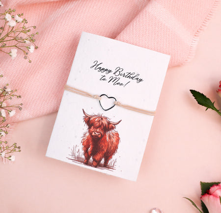 Letterbox Love Happy Birthday bracelet with a silver heart charm, set on a card with a 'Happy Birthday to Moo!' message and Highland cow illustration. Cute and thoughtful birthday gift featuring eco-friendly seed paper design.