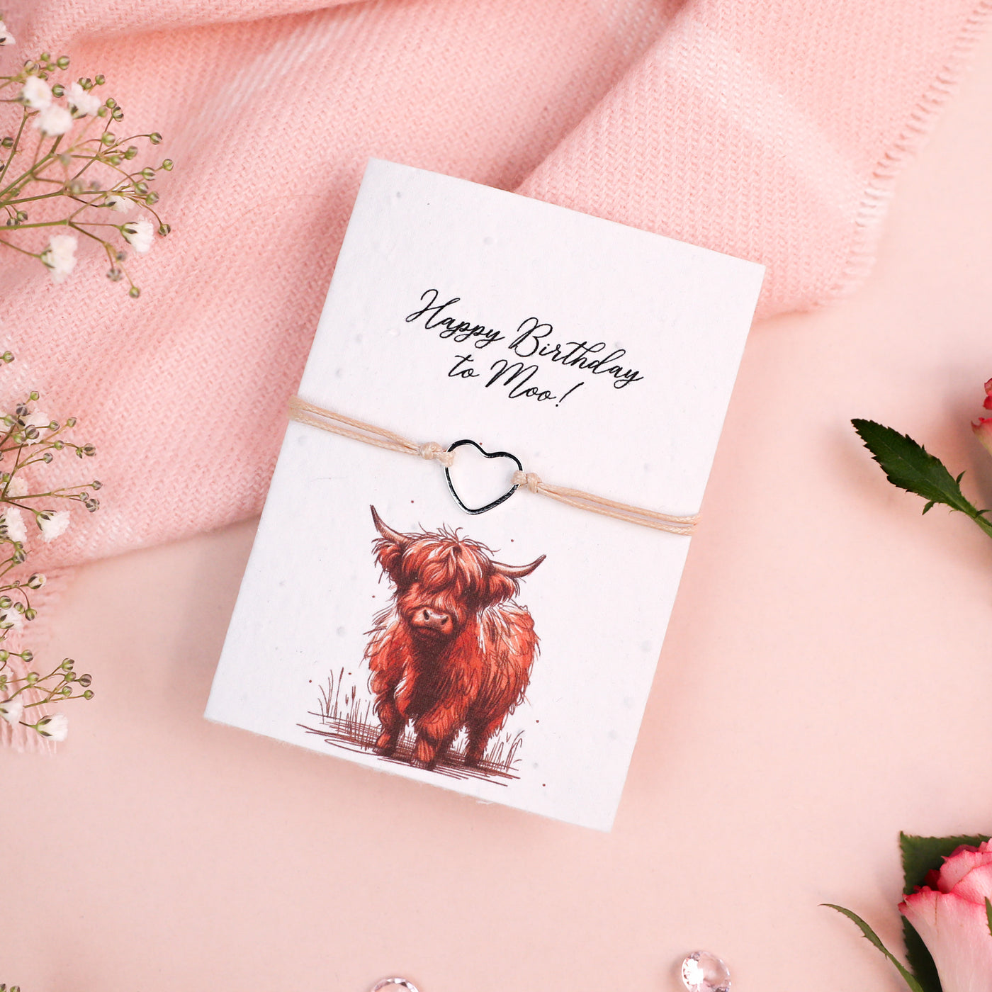 Letterbox Love Happy Birthday bracelet with a silver heart charm, set on a card with a 'Happy Birthday to Moo!' message and Highland cow illustration. Cute and thoughtful birthday gift featuring eco-friendly seed paper design.