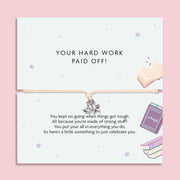 A square greeting card with a thin cord bracelet across the center, featuring a small silver crescent moon and star charm. The top half reads 'YOUR HARD WORK PAID OFF!' and the bottom half has a celebratory message with pastel illustrations of an open book, a notebook, and stacked books on a pink background.