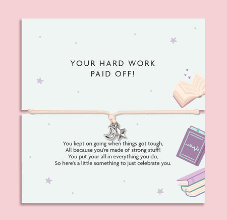 A square greeting card with a thin cord bracelet across the center, featuring a small silver crescent moon and star charm. The top half reads 'YOUR HARD WORK PAID OFF!' and the bottom half has a celebratory message with pastel illustrations of an open book, a notebook, and stacked books on a pink background.