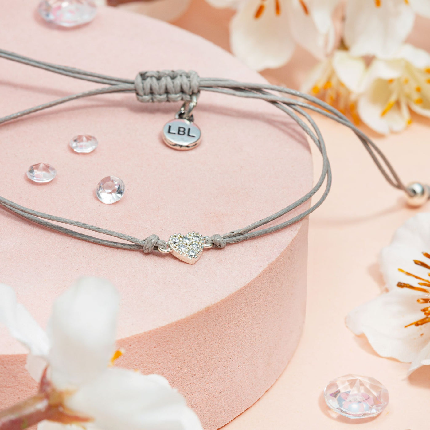 Grey bracelet with heart charm and clear stones on a pink surface with white flowers