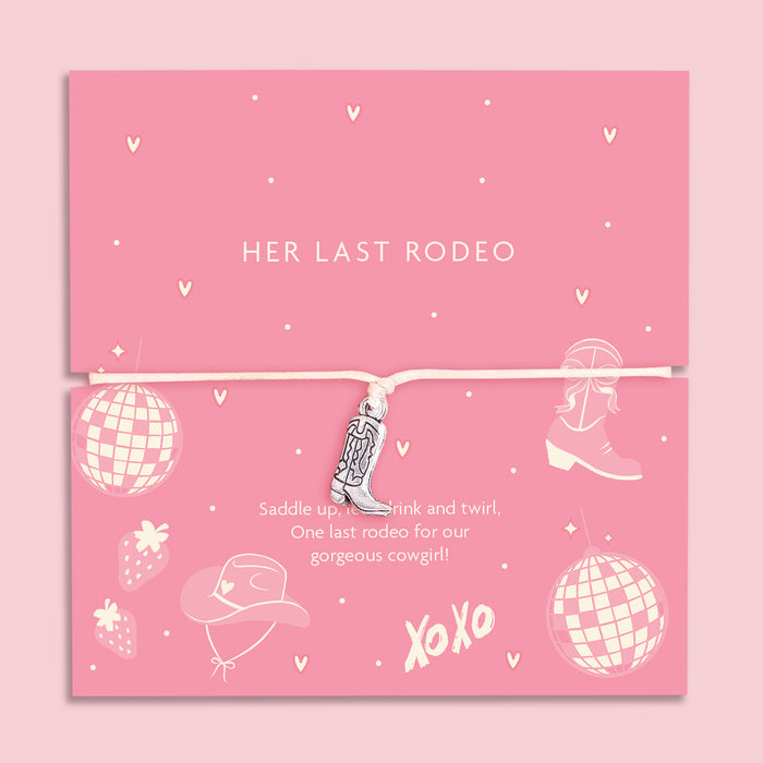 A pink greeting card with the words "HER LAST RODEO" in white uppercase letters. The card is decorated with fun cowgirl-themed illustrations, including disco balls, cowboy boots, a hat, strawberries, and small hearts. A delicate silver cowboy boot charm bracelet on a soft beige cord is wrapped around the card. Below, the message reads: "Saddle up, take a drink, and twirl. One last rodeo for our gorgeous cowgirl!" The background is a soft pink.