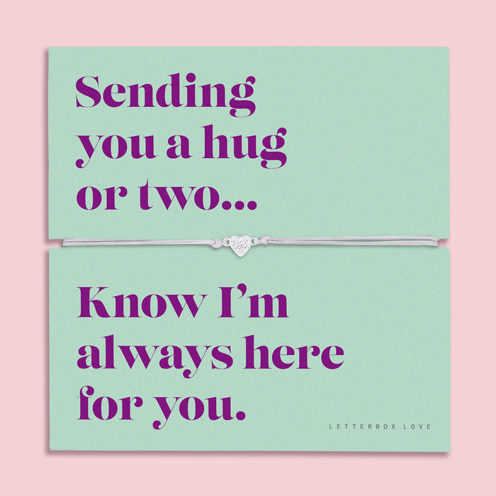A teal-colored bracelet card titled "Always Here for You" from Letterbox Love, featuring a small heart charm on a grey string bracelet. The card includes the message: "Sending you a hug or two... Know I’m always here for you." The setting is a soft pink background with small purple flowers and decorative gems scattered around.