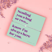 A teal-colored bracelet card titled "Always Here for You" from Letterbox Love, featuring a small heart charm on a grey string bracelet. The card includes the message: "Sending you a hug or two... Know I’m always here for you." The setting is a soft pink background with small purple flowers and decorative gems scattered around.