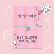A silver beaded bracelet with a heart and key charm displayed on a pink striped gift card that reads 'Hip Hip Hooray' and 'LET'S CELEBRATE YOUR BIG DAY!'. The card is decorated with hand-drawn doodles of stars, hearts, a balloon, and a rainbow, set against a solid pink background with a 'Mini Moments' logo in the top right corner.