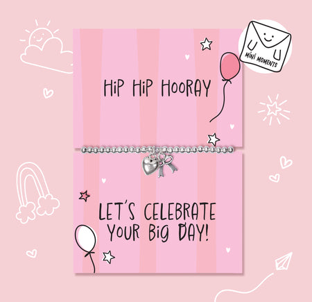 A silver beaded bracelet with a heart and key charm displayed on a pink striped gift card that reads 'Hip Hip Hooray' and 'LET'S CELEBRATE YOUR BIG DAY!'. The card is decorated with hand-drawn doodles of stars, hearts, a balloon, and a rainbow, set against a solid pink background with a 'Mini Moments' logo in the top right corner.