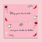Adjustable string bracelet with small silver book charm presented on pink card reading “May Your Tea Be Hot and Your Books Be Hotter” with illustrated teapots, books and red chillies.