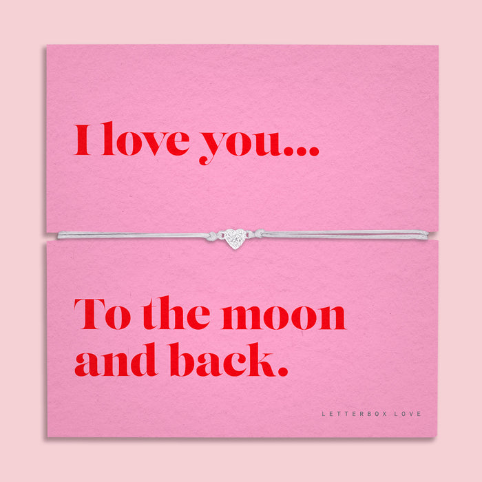 A pink bracelet card titled "I love you to the moon and back" from Letterbox Love, featuring a small heart charm on a grey string bracelet. The setting is a soft pink background with small pink flowers and decorative gems scattered around.