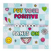 An image of a motivational card featuring a beaded bracelet with white letter beads spelling "YOU CAN." The card has a light blue checkered background with vibrant text that reads: "Put your positive pants on." The design includes playful illustrations of smiley faces, hearts, lightning bolts, and a cartoon image of polka-dotted underwear. The card is decorated with bright colors and fun graphics, creating a lighthearted and positive message. A playful gift designed to inspire confidence and a positive mind