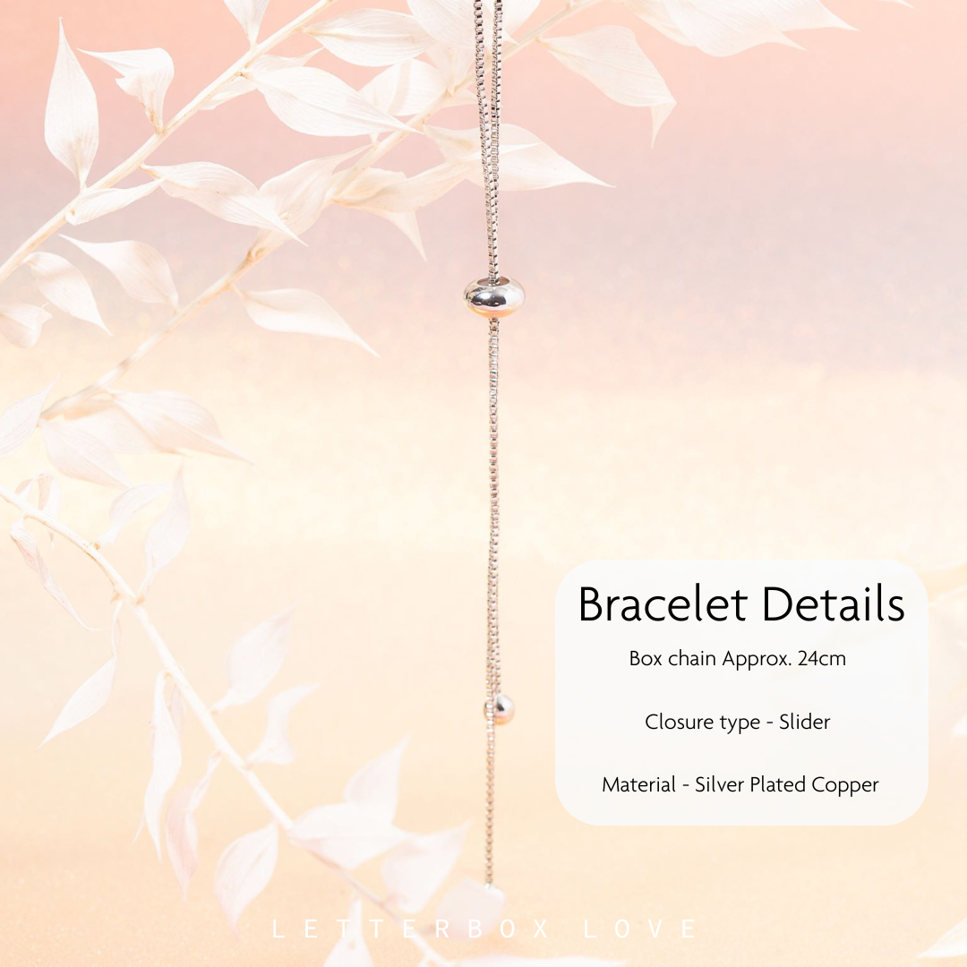 Silver-plated copper box chain bracelet with a sliding closure and small bead accents, shown hanging against a soft peach background with white leaf branches and text detailing the product features.