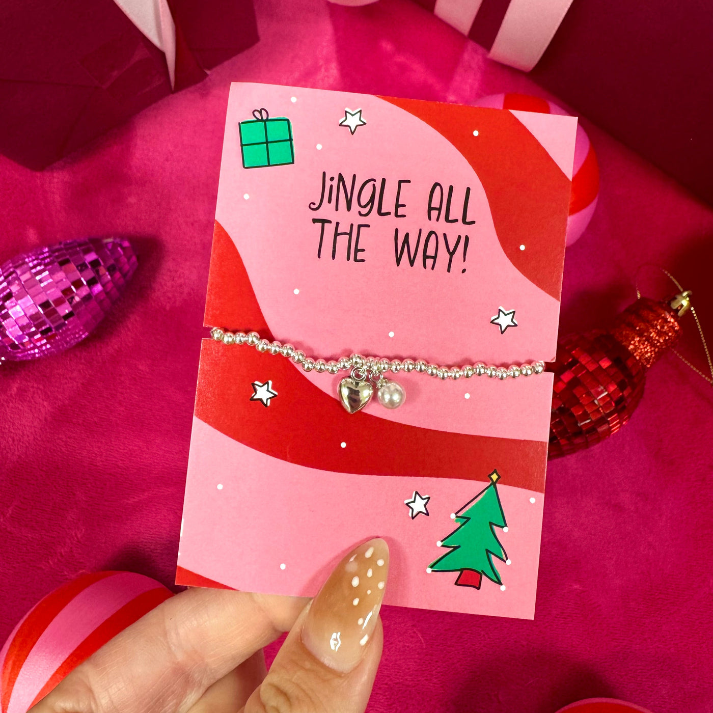 A silver beaded bracelet with a small heart and star charm is displayed on a pink Christmas-themed gift card held by a hand. The card is surrounded by festive decorations, including red gift boxes with white ribbons, a purple ornament, and a red ornament, on a deep pink background.
