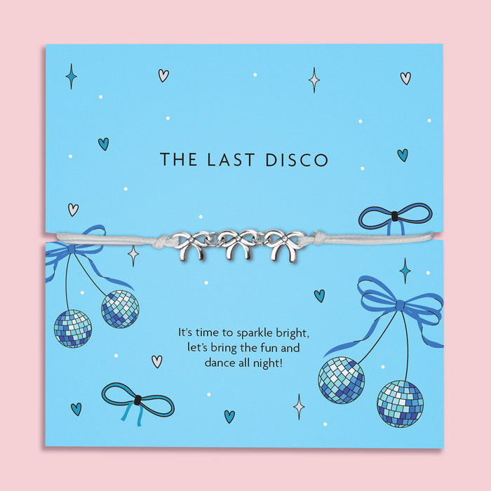 A vibrant blue card with the words "THE LAST DISCO" in black uppercase letters. The card is decorated with fun disco-themed illustrations, including sparkling disco balls, blue bows, hearts, and stars. A delicate bracelet with three silver bow charms on a soft ivory cord is wrapped around the card. Below, the message reads: "It’s time to sparkle bright, let’s bring the fun and dance all night!" The background is plain white, highlighting the bracelet and card design.