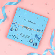 A vibrant blue card with the words "THE LAST DISCO" in black uppercase letters. The card is decorated with fun disco-themed illustrations, including sparkling disco balls, blue bows, hearts, and stars. A delicate bracelet with three silver bow charms on a soft ivory cord is wrapped around the card. Below, the message reads: "It’s time to sparkle bright, let’s bring the fun and dance all night!" The background is plain white, highlighting the bracelet and card design.