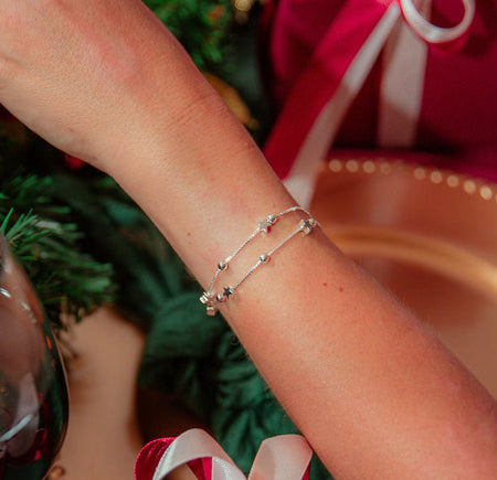 A delicate silver double-strand bracelet with small star charms worn on a person's wrist, set against a festive Christmas background with wrapped gifts and greenery.