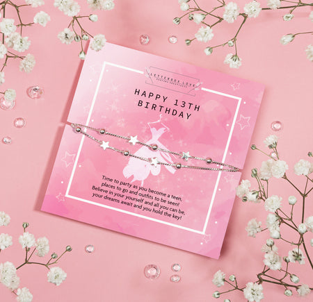 A pink themed 13th birthday card with a 'Happy 13th Birthday' greeting, adorned with silver starry chains and surrounded by white blossoms and gemstones on a pink background. 