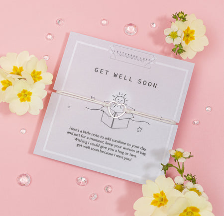 Charming 'Get Well Soon' card on a soft pink background, tied with a delicate white ribbon. The card features a cheerful sun illustration and a comforting message. Beside the card are vibrant yellow flowers with white petals and scattered clear gemstones, evoking a sense of warmth and well-wishes.